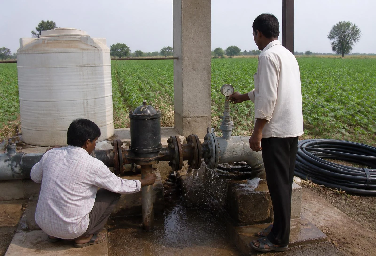 Two farm staff at a pump house during a follow-up check, one adjusting a valve on the mainline while the other reads a pressure gauge.